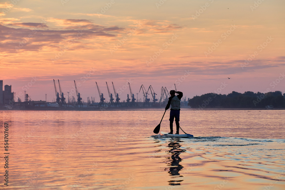 Orange winter sunset and silhouette of boy rowing on supboards (SUP) at calm Danube river at the morning against port cranes