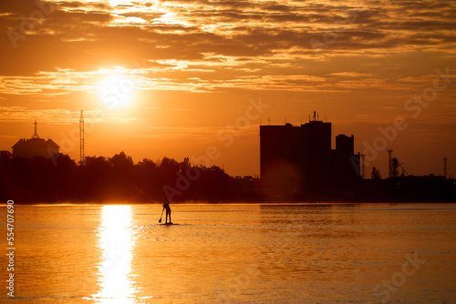 Silhouette of a woman paddle on an inflatable SUP board at sunrise and paddling through the shiny water surface of river