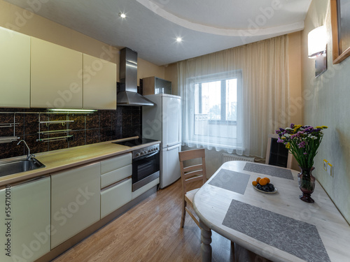 Modern interior of kitchen in apartment. Table and chairs. Fridge  oven and fan. Window with tulle.