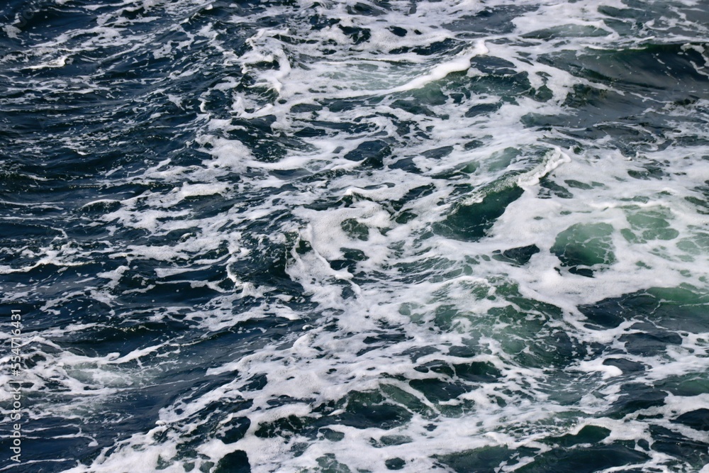 Stern waves with white foam tips on greyish blue sea water, photo taken from aboard ship. Selective focus