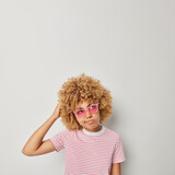 Photo of clueless curly haired woman scratches head looks doubtful thinks about making decision wears trendy sunglasses and casual striped t shirt isolated over grey background. Hmm let me think