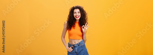 Charming young charistmatic woman with curly hairstyle in cropped top and shorts showing okay or approval gesture smiling broadly liking awesome idea of friend where hang out over orange wall photo