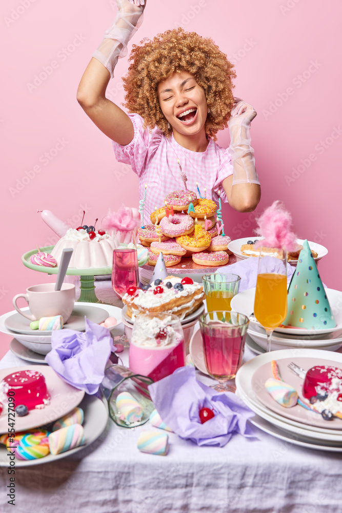 Happy curly haired woman dances carefree near festive table with diffrent desserts celebrates birthday wears dress and silk gloves keeps eyes closed isolated over pink background. Celebration concept