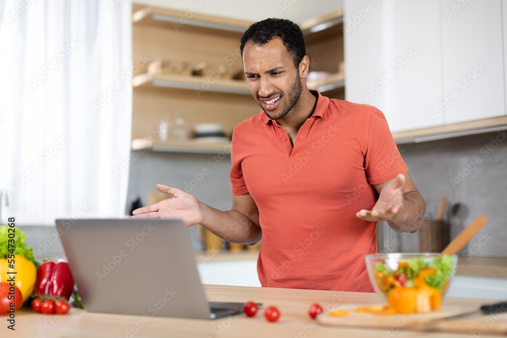 Upset angry young black man in red t-shirt gesturing with hands at computer, has problems with service Upset angry young black man in red t-shirt gesturing with hands at computer, has problems with service