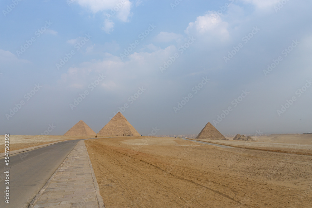 The three great pyramids of the Giza complex, seen from the back, with the yellow sand, a road next to the pyramids and the sky with clouds.