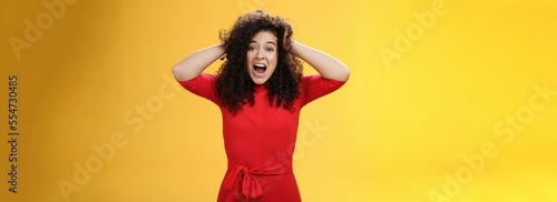 Girl feeling pressure standing anxious in panic holding hands on curly hair yelling at camera disturbed, freaked out being tensed and upset with bad situation, standing troubled over yellow background photo