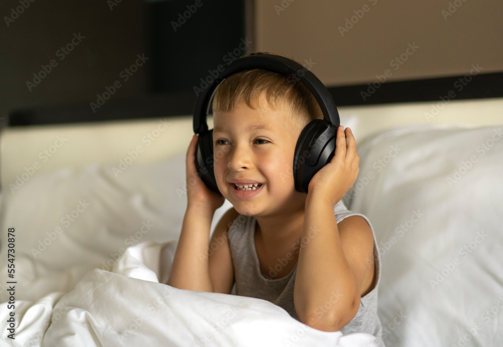 Little sleepy boy sitting in bed wearing wireless headphones and listening to music Little sleepy boy sitting in bed wearing wireless headphones and listening to music