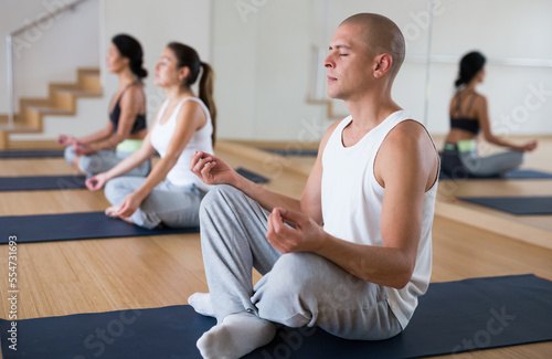 Focused man meditating in yoga position Padmasana during group training at gym