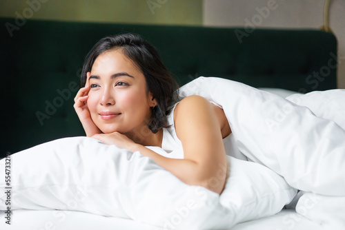 Dreamy asian girl lying in her bed in morning, covered with warm duver and leaning on white pillow, looking at window and thinking photo