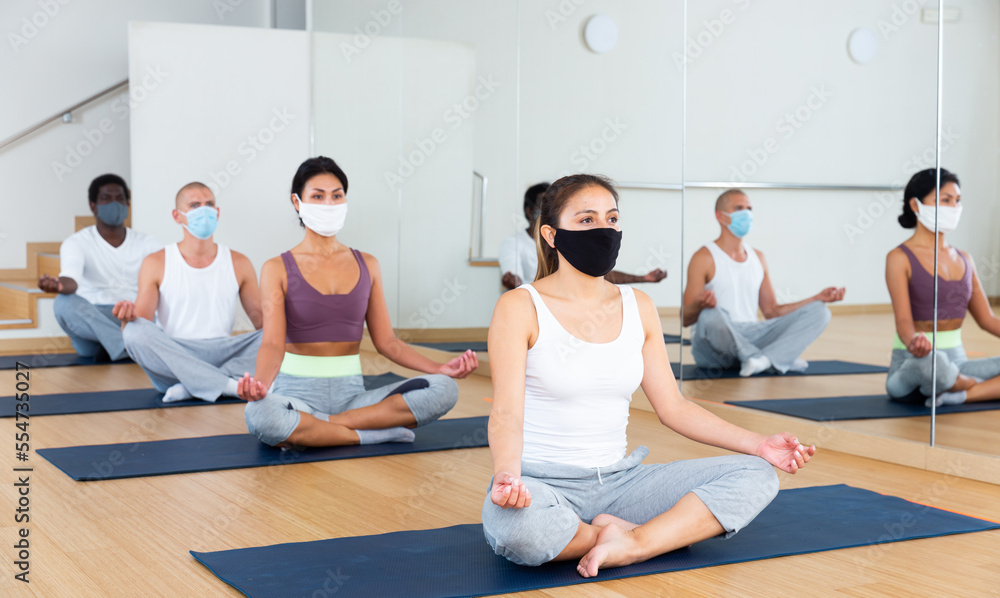 Multiethnic group of sporty people wearing protective masks to prevent viral infections, practicing yoga poses while exercising at fitness center
