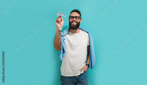Young handsome hipster man with a beard holds a paper airplane in his hand, smiling happily.