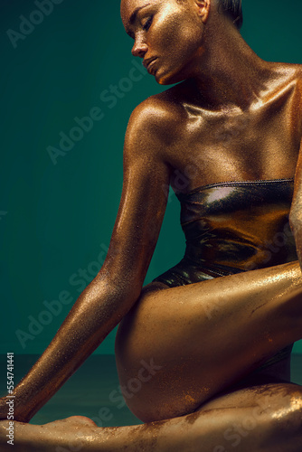 Model girl with bright golden sparkles on her body posing, full length portrait of beautiful sexy woman with glowing body skin. Swimsuit. Art design make up. Glitter gold sequins on skin body art
