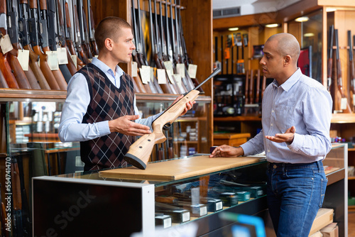 Confident experienced salesperson helping Hispanic customer choose right hunting rifle at gun store photo