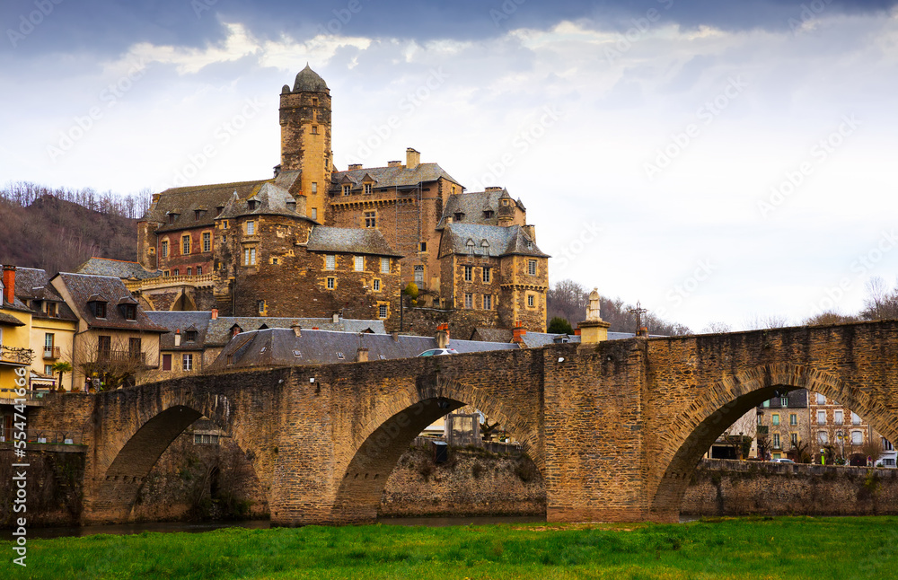View of medieval castle of Chateau Estaing and old arched bridge across river Lot in cloudy winter day, Aveyron, France..