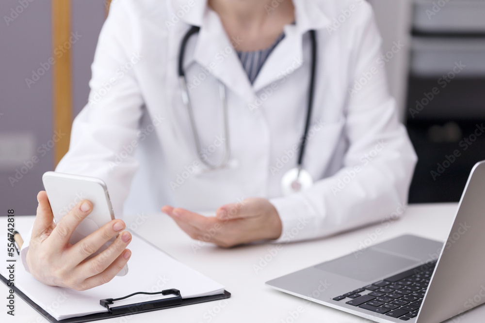 A doctor in a cubicle at a white table is talking on a video link holding a phone in his hand. In the doctor's office, there is a laptop on a white table and a tablet for notes on paper. High quality A doctor in a cubicle at a white table is talking on a video link holding a phone in his hand. In the doctor's office, there is a laptop on a white table and a tablet for notes on paper. High quality