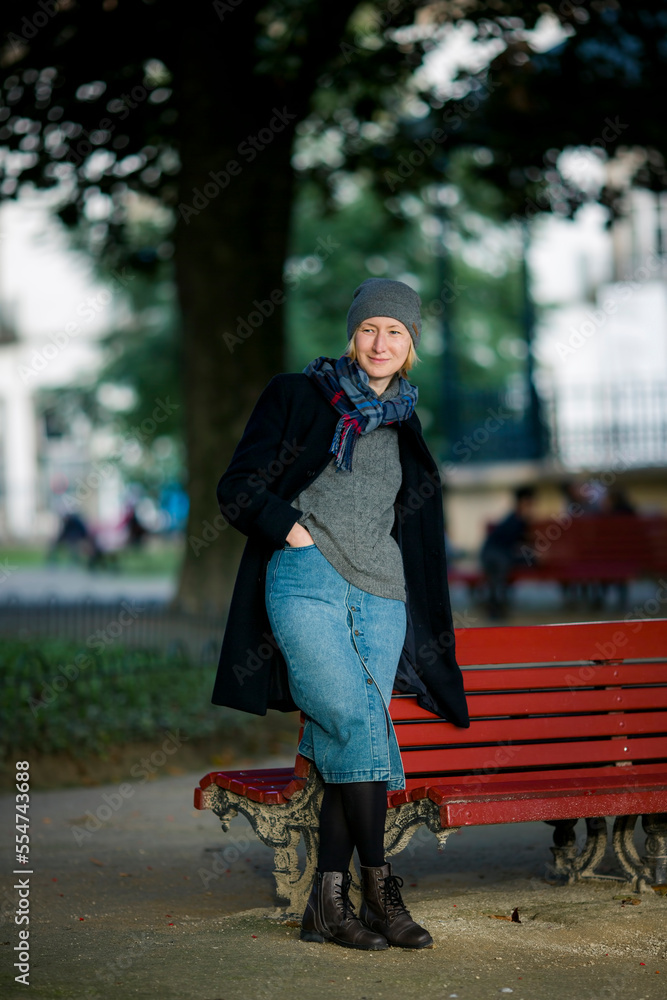 A woman in an autumn coat stands in the park near a wooden bench.