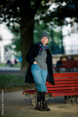 A woman in an autumn coat stands in the park near a wooden bench.
