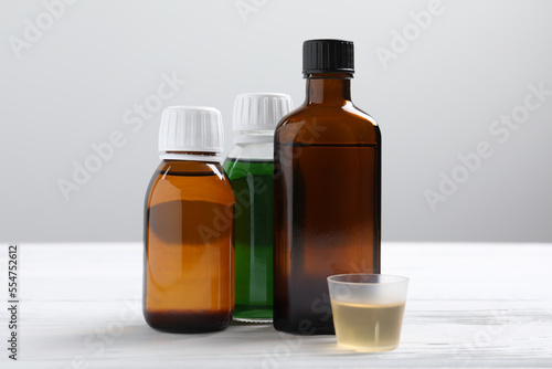 Bottles of syrup and measuring cup on white table against light grey background. Cold medicine