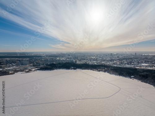 Snow-covered forest on lake shore with ice at sunset and the city on horizon, auerial view © Dmitrii Potashkin