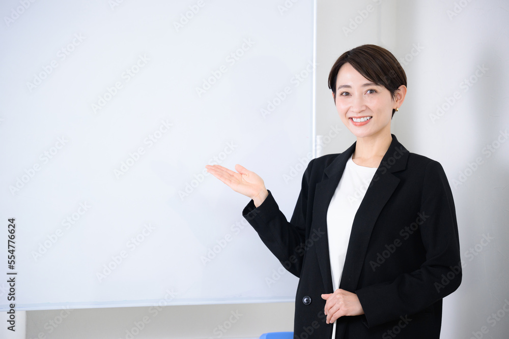 スーツ姿でオススメのポーズをする使いやすいビジネス系女性の黒スーツのイメージ　ホワイトボード付き　カメラ目線