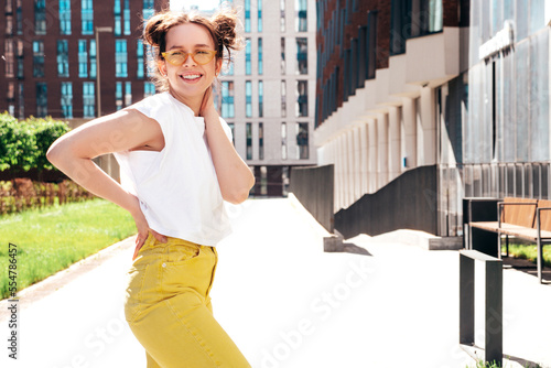 Young beautiful smiling hipster woman in trendy summer yellow jeans clothes. Carefree teen model posing in the street at sunset. Positive female outdoors. Cheerful and happy. In sunglasses