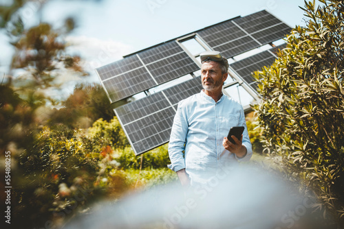 Smiling mature man standing with smart phone in front of solar panels photo