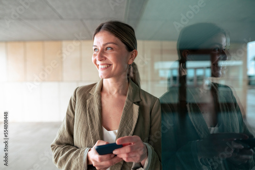 Happy businesswoman with smart phone leaning on glass wall photo