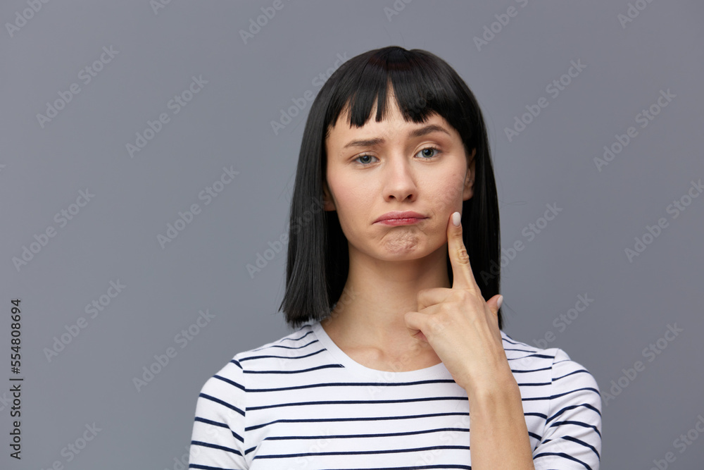 a sweet, attractive, thoughtful woman stands on a gray background in a white striped T-shirt and holds her index finger near her face. Horizontal photo a sweet, attractive, thoughtful woman stands on a gray background in a white striped T-shirt and holds her index finger near her face. Horizontal photo