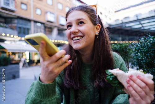 Smiling young woman holding sandwich talking through speaker of mobile phone photo