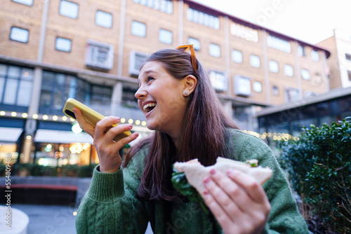Happy woman holding sandwich talking through speaker of mobile phone in front of building photo
