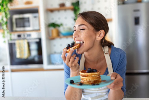 Young woman making pancakes at kitchen. Young housewife enjoying blueberry pancakes for breakfast