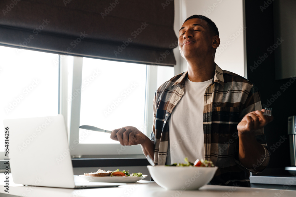 Young pleased african man using laptop while having lunch at home Young pleased african man using laptop while having lunch at home