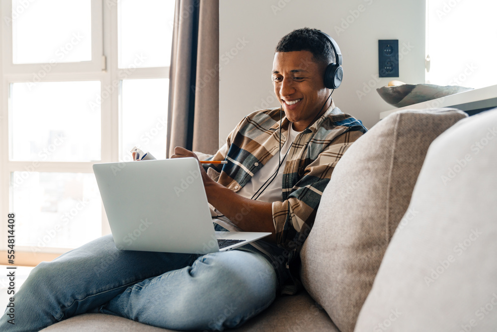 Young african man studying on laptop and writing down notes while sitting on couch Young african man studying on laptop and writing down notes while sitting on couch