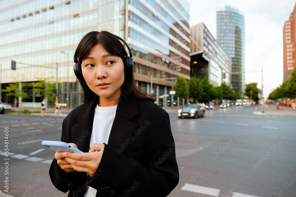 Fototapeta premium Young woman using cellphone and headphones while walking on street