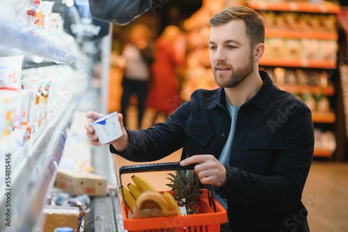 Customer In Supermarket. Man Doing Grocery Shopping Standing With Cart Choosing Food Product Indoors. Guy Buying Groceries In Food Store. Selective Focus, Copy Space.