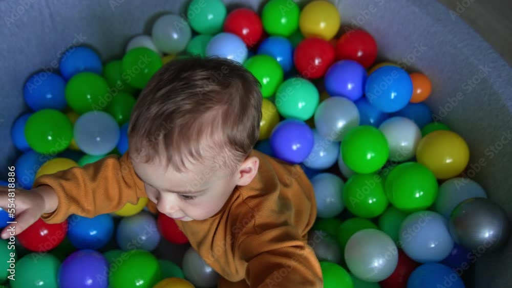 Adorable kid sitting in dry pool with balls looks up curiously into camera. Lovely boy wants to stand up but fails. Top view. Adorable kid sitting in dry pool with balls looks up curiously into camera. Lovely boy wants to stand up but fails. Top view.