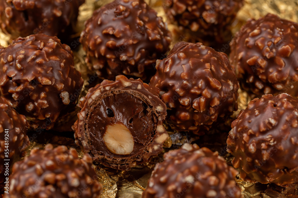 Chocolate and hazelnut ball candies. Whole roasted hazelnut in a thin wafer shell filled with hazelnut chocolate, covered milk chocolate chopped hazelnuts. Confectionery on a gold wrapping foil. Chocolate and hazelnut ball candies. Whole roasted hazelnut in a thin wafer shell filled with hazelnut chocolate, covered milk chocolate chopped hazelnuts. Confectionery on a gold wrapping foil.