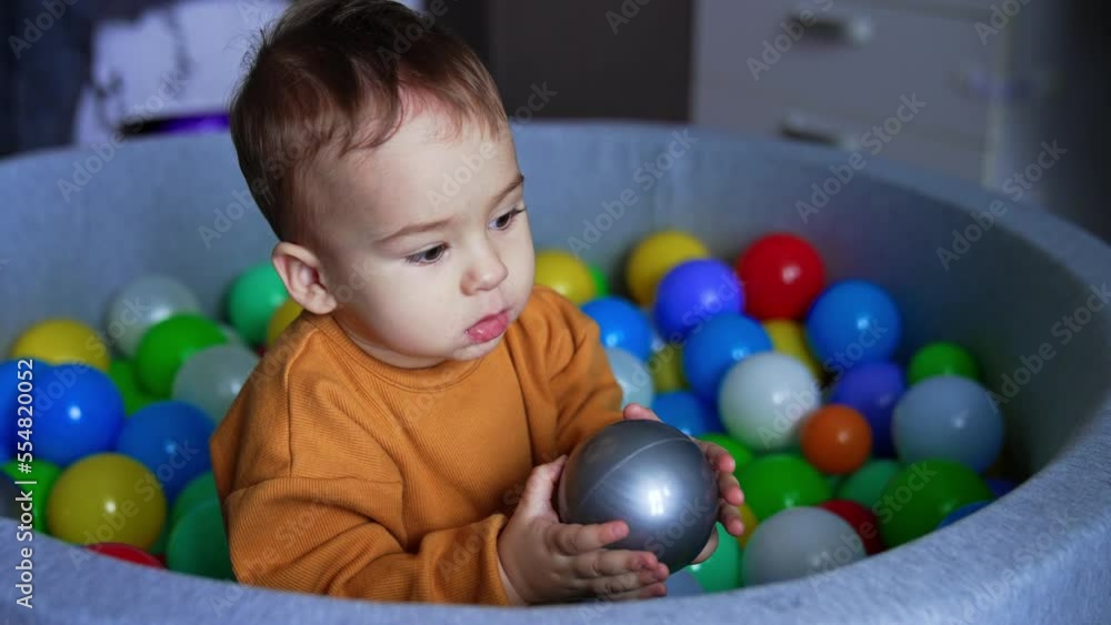 Beautiful Caucasian baby sitting calm holding a grey ball in hands. Kid is in the dry pool and throws the ball out of the basin. Close up. Beautiful Caucasian baby sitting calm holding a grey ball in hands. Kid is in the dry pool and throws the ball out of the basin. Close up.