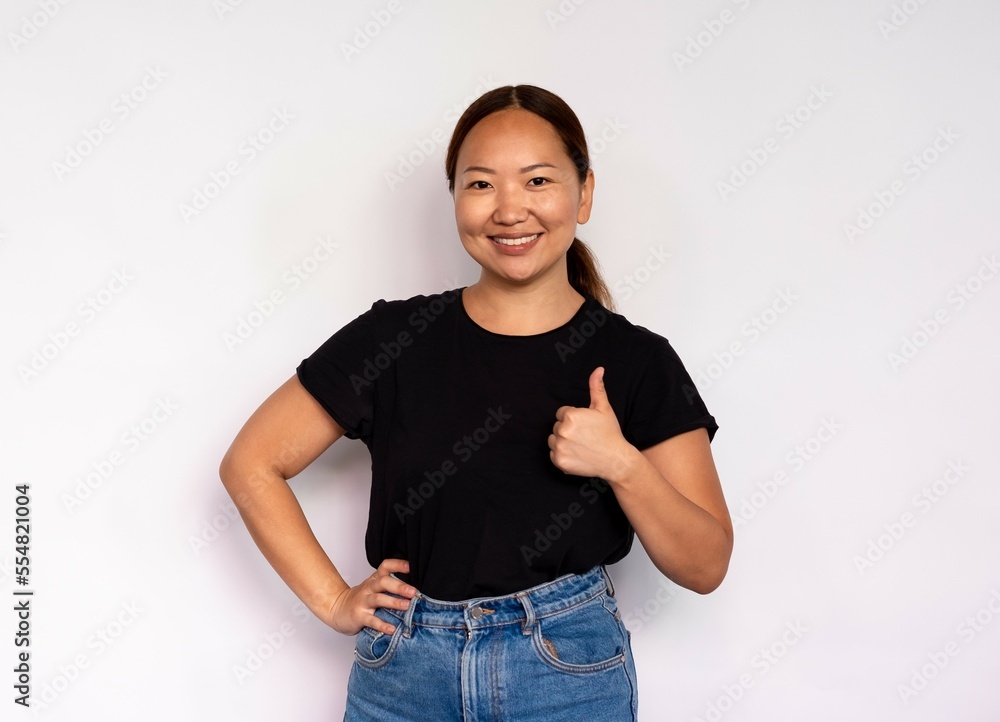 Portrait of cheerful young woman making thumb up over white background. Asian lady wearing black T-shirt and jeans looking at camera and smiling. Approval concept Portrait of cheerful young woman making thumb up over white background. Asian lady wearing black T-shirt and jeans looking at camera and smiling. Approval concept