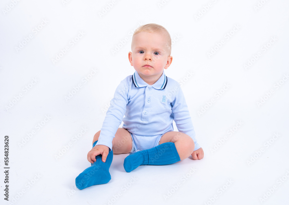 Beautiful blond baby boy wearing blue sitting against white background, having serious thoughtful look, smiling and looking to the camera. Beautiful blond baby boy wearing blue sitting against white background, having serious thoughtful look, smiling and looking to the camera.
