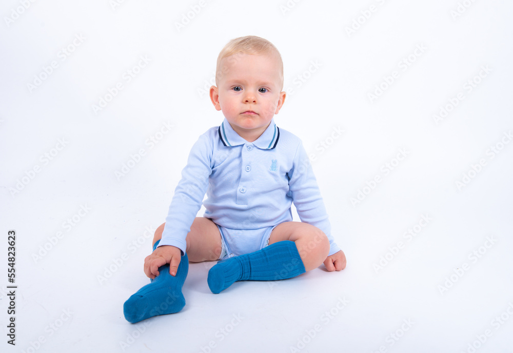 Beautiful blond baby boy wearing blue sitting against white background, having serious thoughtful look, smiling and looking to the camera. Beautiful blond baby boy wearing blue sitting against white background, having serious thoughtful look, smiling and looking to the camera.