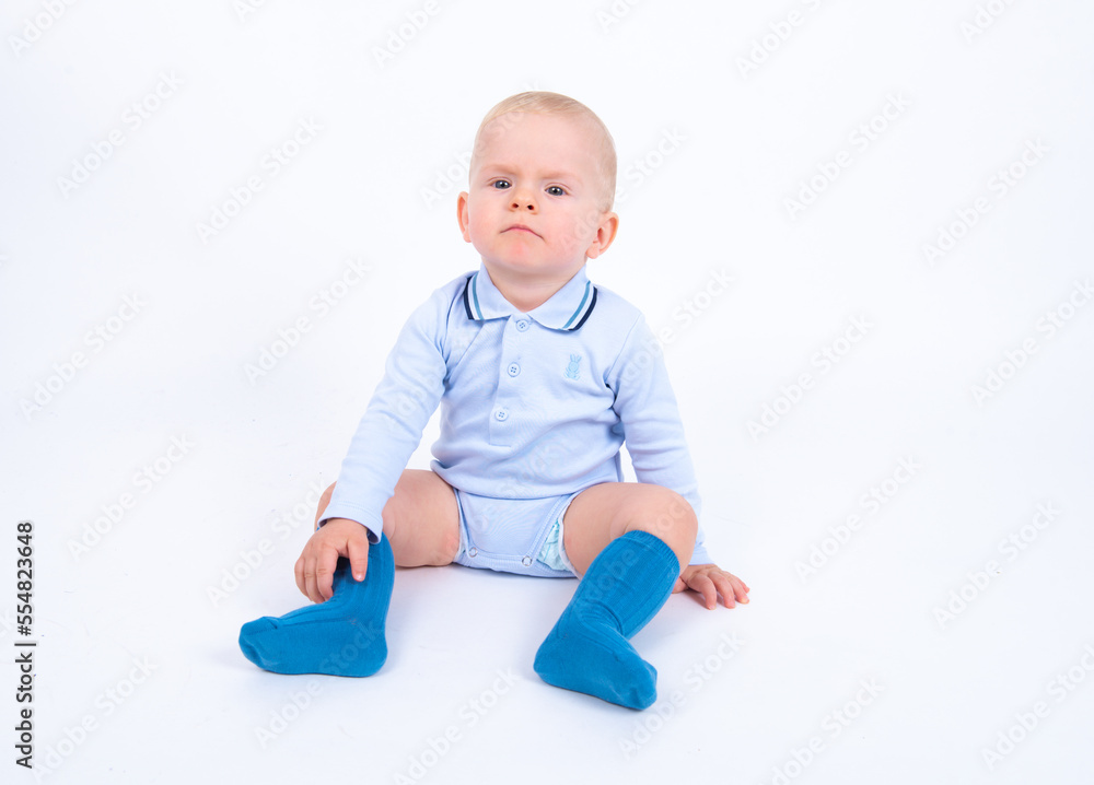 Beautiful blond baby boy wearing blue sitting against white background feeling angry and looking to the camera. Beautiful blond baby boy wearing blue sitting against white background feeling angry and looking to the camera.