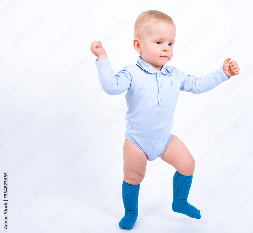 Beautiful blond baby boy wearing blue standing against white background smiling and looking to the camera. Baby taking his first steps. Beautiful blond baby boy wearing blue standing against white background smiling and looking to the camera. Baby taking his first steps.