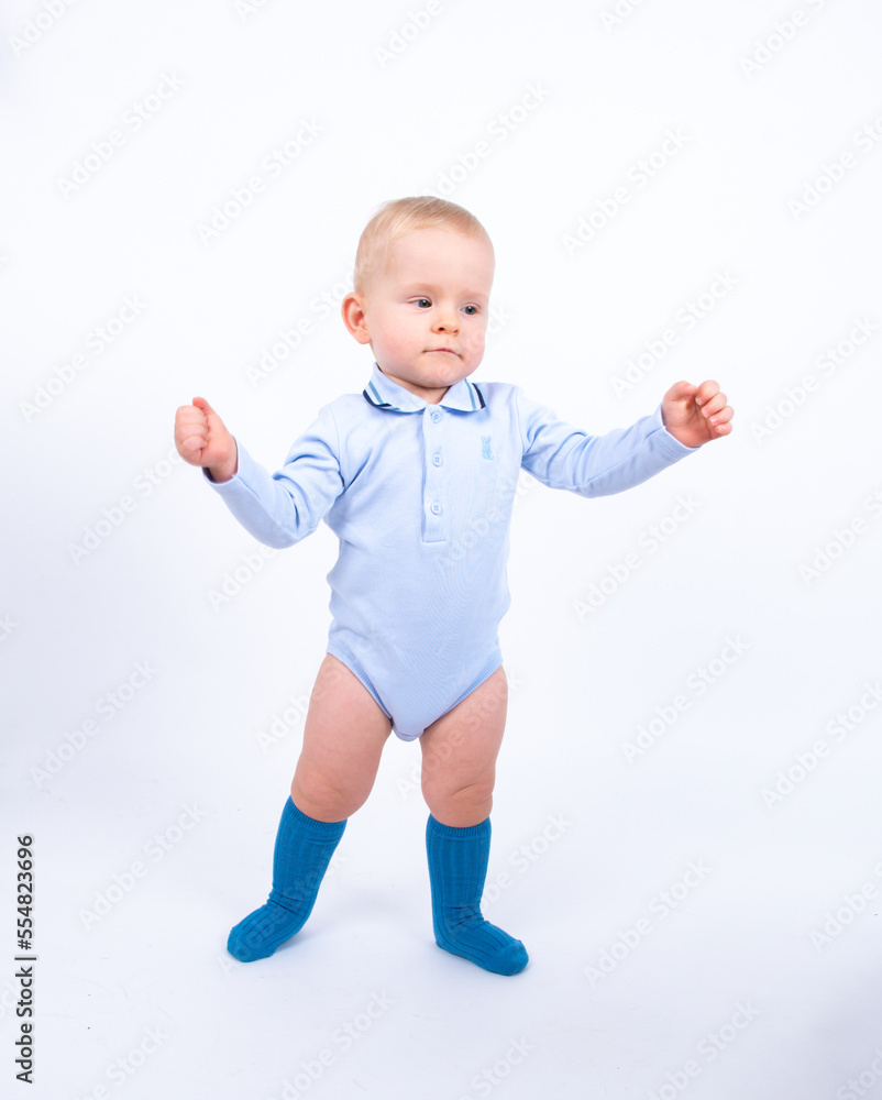 Beautiful blond baby boy wearing blue standing against white background smiling and looking to the camera. Beautiful blond baby boy wearing blue standing against white background smiling and looking to the camera.