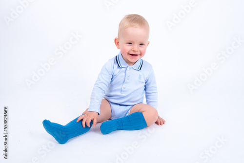 Beautiful blond baby boy wearing blue sitting against white background smiling and looking to the camera. 