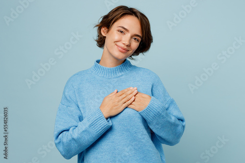 Young greatful happy fun smiling caucasian woman in knitted sweater look camera put folded hands on heart isolated on plain pastel light blue cyan background studio portrait People lifestyle concept
