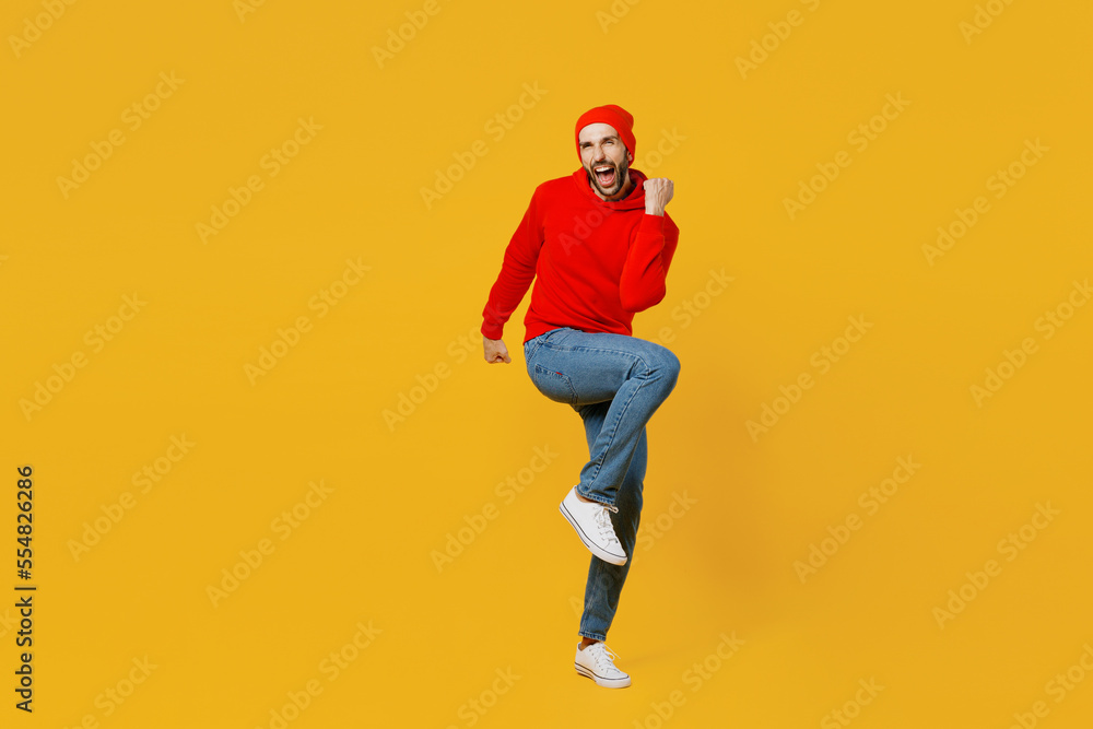 Full body young happy fun caucasian man wear red hoody hat look camera doing winner gesture raise up leg clench fist isolated on plain yellow color background studio portrait People lifestyle concept Full body young happy fun caucasian man wear red hoody hat look camera doing winner gesture raise up leg clench fist isolated on plain yellow color background studio portrait People lifestyle concept