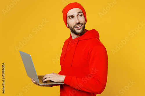 Side view young smart IT man wears red hoody hat hold use work on laptop pc computer look aside on workspace area isolated on plain yellow color background studio portrait. People lifestyle concept. © ViDi Studio