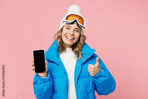 Snowboarder woman wear blue suit goggles mask hat ski jacket hold mobile cell phone blank screen show thumb up isolated on plain pastel pink background. Winter extreme sport hobby trip relax concept.