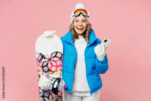 Snowboarder smiling woman wear blue suit goggles mask hat ski padded jacket hold use mobile cell phone isolated on plain pastel pink background. Winter extreme sport hobby weekend trip relax concept.